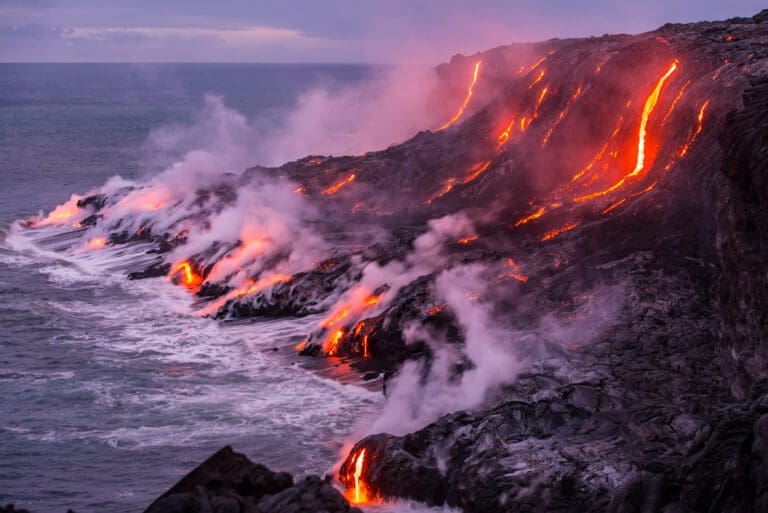 Lava pouring on the side of a mountain into the ocean.